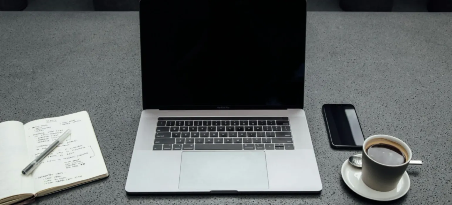 Turned-off MacBook Pro between cup of coffee, iPhone, notebook, and pen