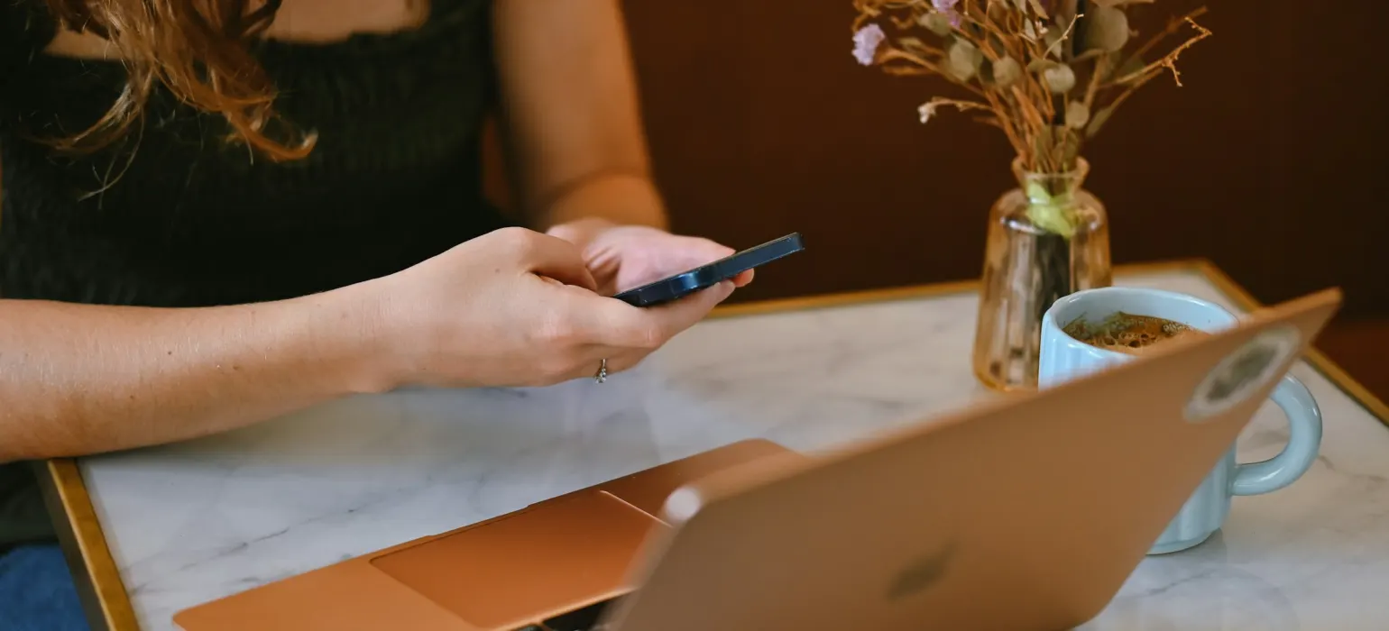 A person using a smartphone at a marble table with a laptop and flowers