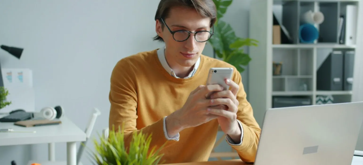 Man in glasses using phone at desk with laptop